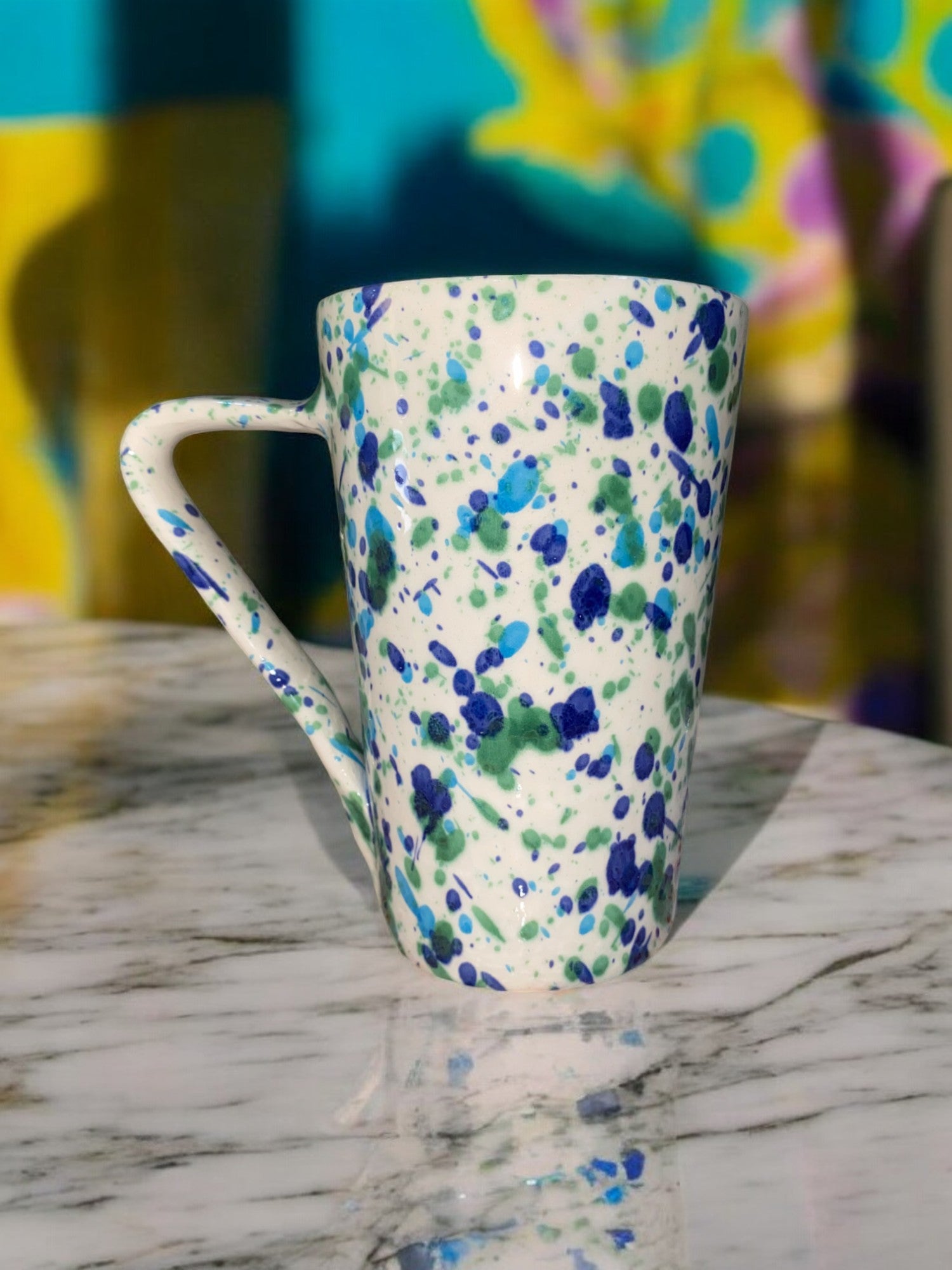 Grand mug artisanal en céramique, de forme ronde, avec un motif moucheté bleu et vert sur un fond blanc. La surface est légèrement texturée, et le mug a une anse large pour une prise en main confortable.