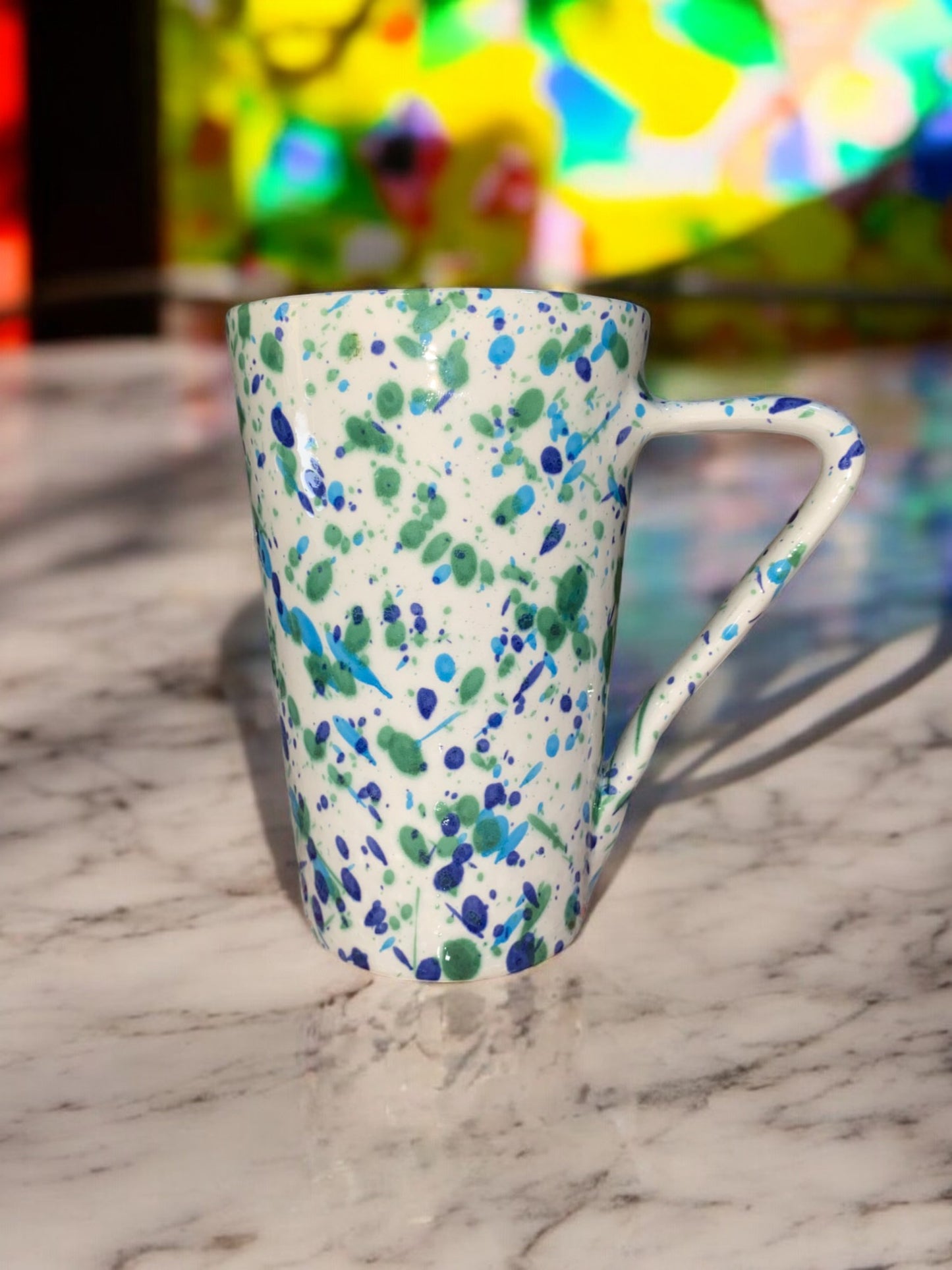 Grand mug artisanal en céramique, de forme ronde, avec un motif moucheté bleu et vert sur un fond blanc. La surface est légèrement texturée, et le mug a une anse large pour une prise en main confortable.